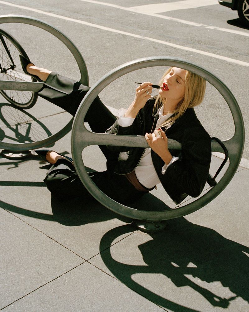 Fashion shot of a blonde adult woman applying red lipstick while reclining on a metal bike rack in sunlight on an urban street.