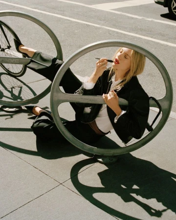 Fashion shot of a blonde adult woman applying red lipstick while reclining on a metal bike rack in sunlight on an urban street.