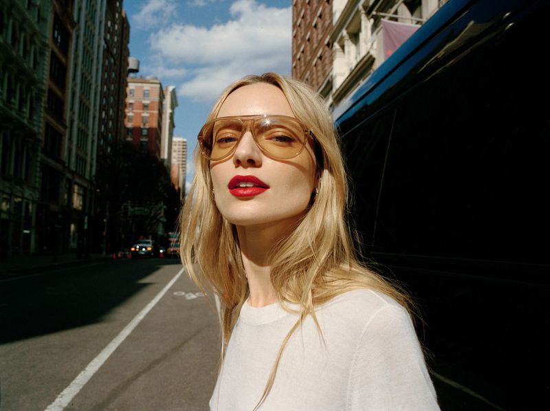 Portrait of a blonde woman in sunglasses and a white sweater standing on a city street with tall buildings in the background.