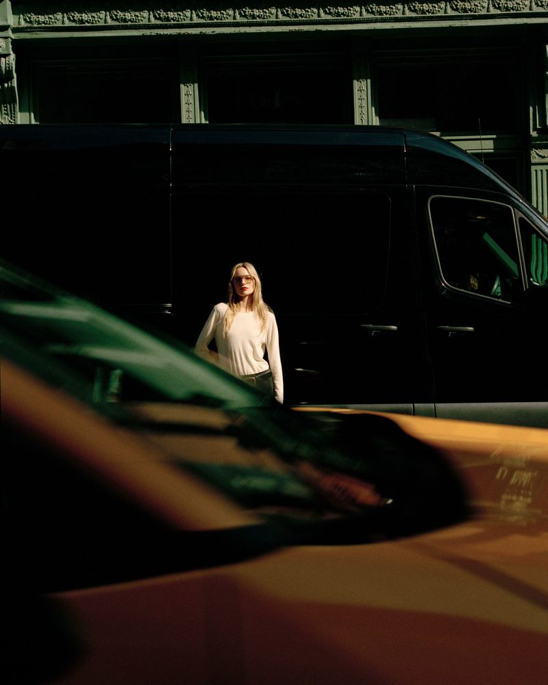 A woman with long blonde hair stands in front of a black van on a city street. She wears sunglasses, a cream-colored sweater, and dark pants.