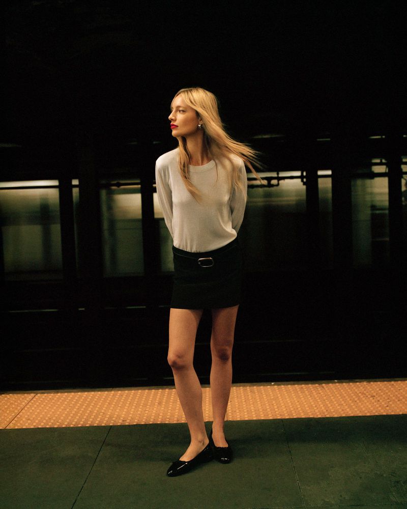 A woman with long blonde hair, wearing a white long-sleeve shirt, and a black skirt, stands on a subway platform next to a train.
