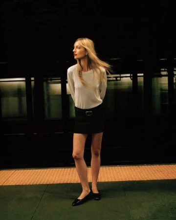 A woman with long blonde hair, wearing a white long-sleeve shirt, and a black skirt, stands on a subway platform next to a train.