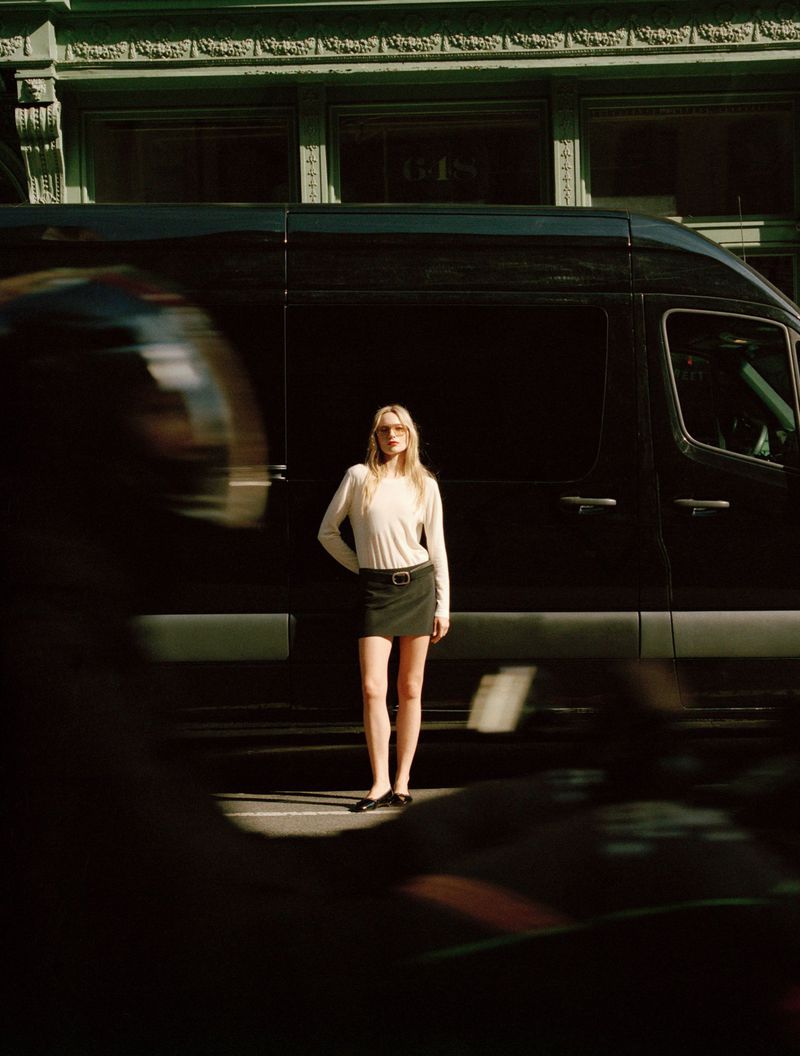 Blonde woman in a skirt and long-sleeve shirt stands near a black van on a city street, partly obscured by blurred motion in the foreground.