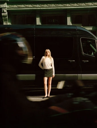 Blonde woman in a skirt and long-sleeve shirt stands near a black van on a city street, partly obscured by blurred motion in the foreground.