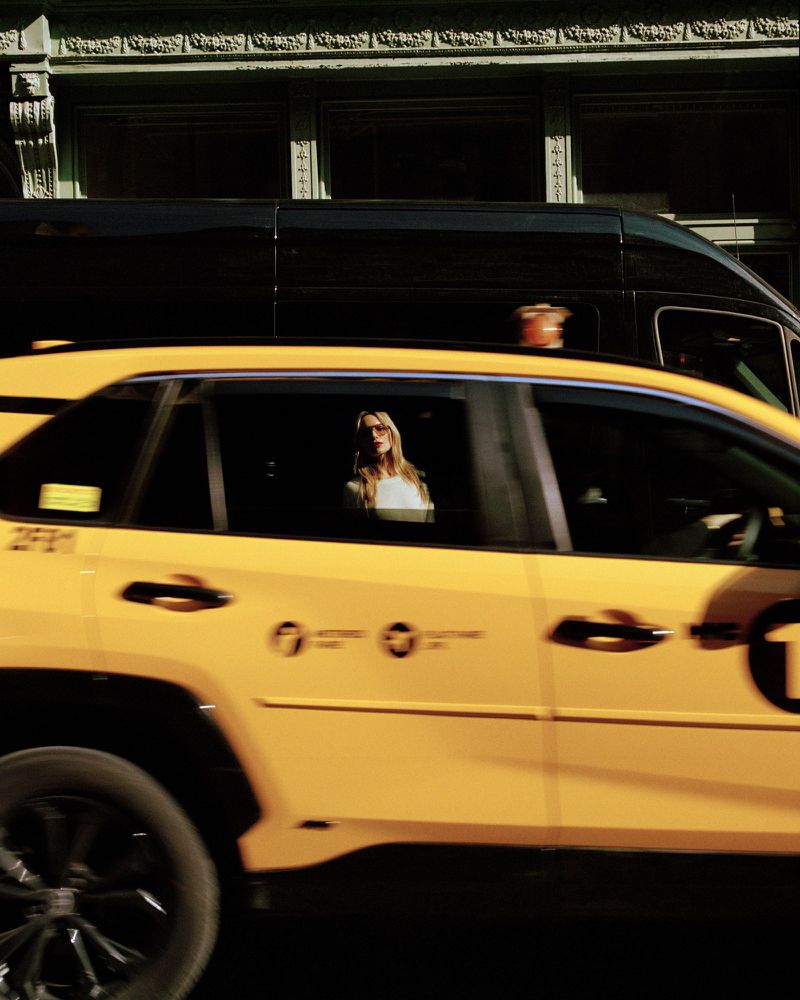 A yellow New York City taxi cab with a passenger is partially visible, with an urban building and a black vehicle in the background.