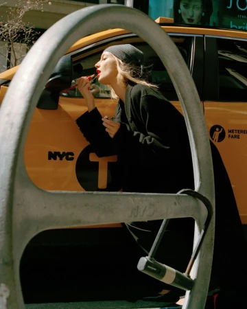 Adult female model applying lipstick on the street, reflecting in a yellow taxi mirror, wearing a black coat and beanie in daylight.