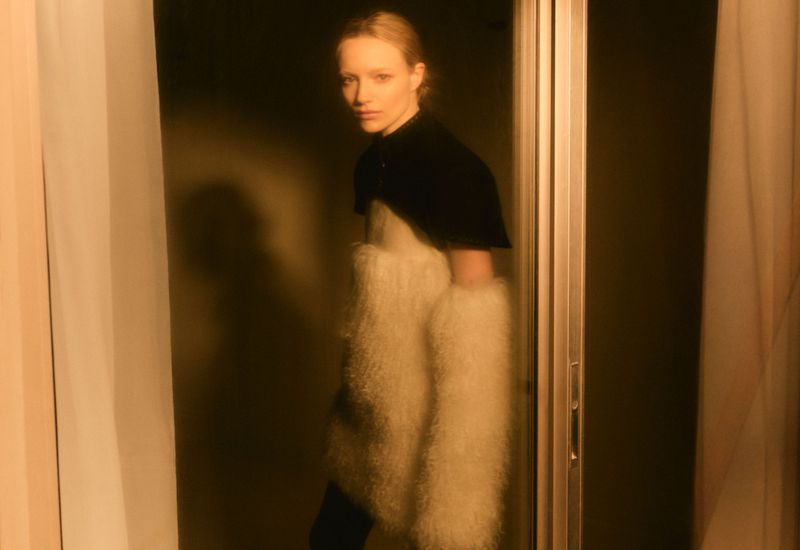 Blonde model in black top and white faux fur sleeves poses in a warmly lit interior setting with sliding doors in a studio-like environment.