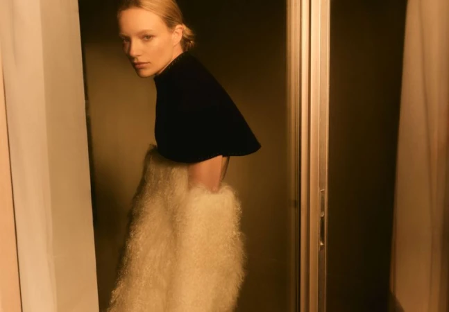 A blonde woman wearing a black top and white shaggy skirt poses in a dimly lit indoor setting, framed by vertical lines and warm light.