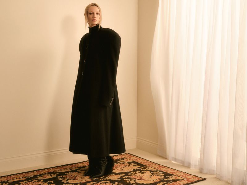 A slender woman in a striking black coat poses in front of sheer curtains indoors, standing on an ornate rug with neutral wall backdrop.