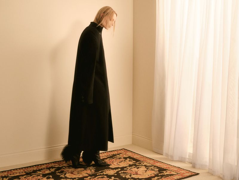 Fashion shot of a young adult female model wearing a black overcoat and boots, posing indoors against a neutral wall and a patterned rug.