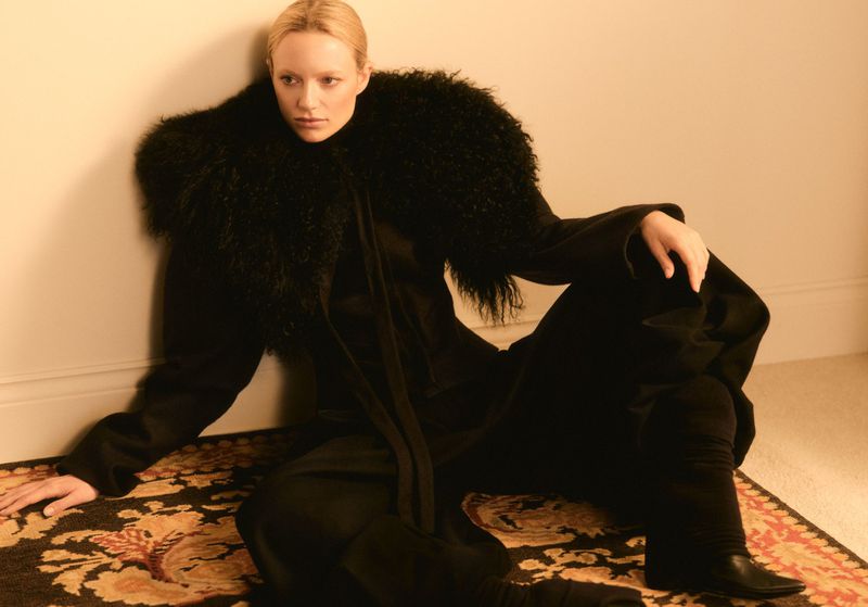 Fashion portrait of a fair-skinned woman with blonde hair sitting on a carpet, wearing a black coat with a large fur collar indoors.
