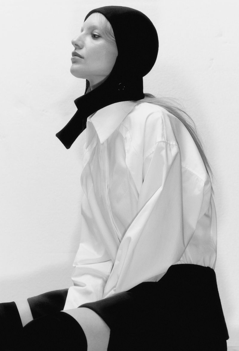 Monochrome fashion portrait of a woman in a black head covering and white collared shirt against a plain white background, studio lighting.