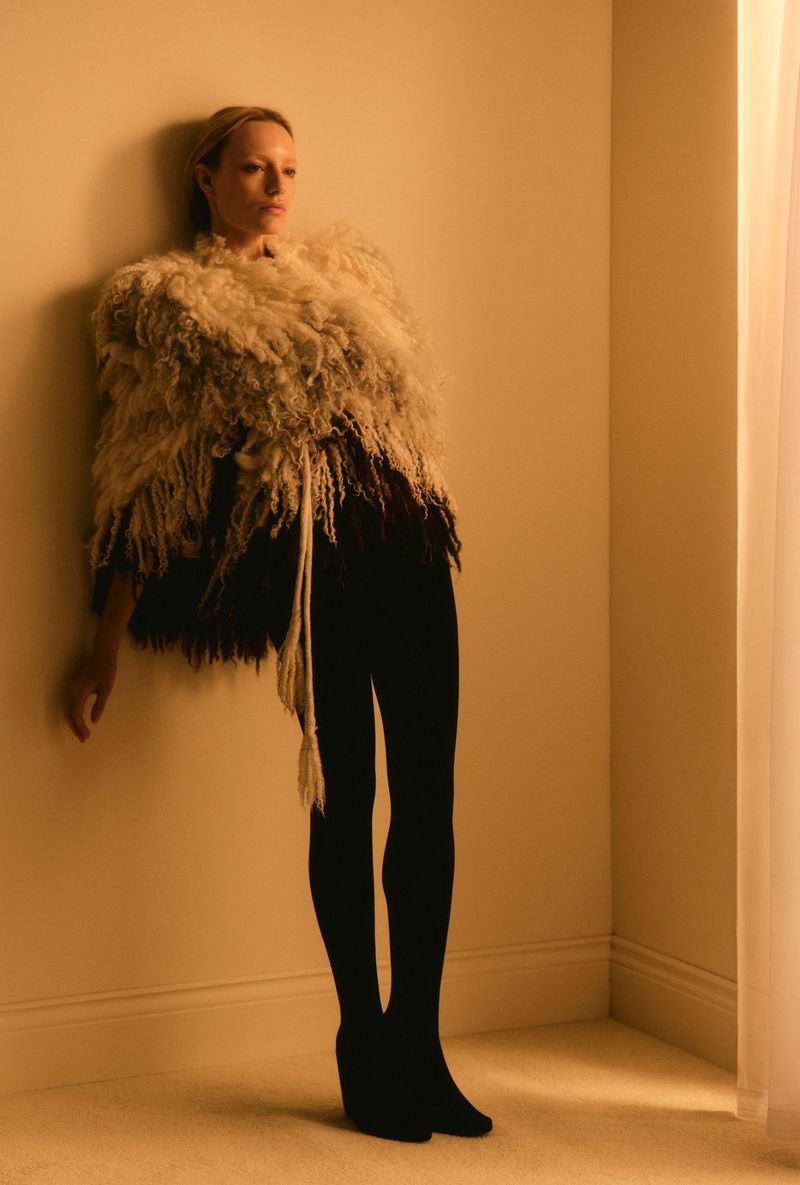 Full-length shot of a woman indoors, leaning against a wall and wearing a fringed shawl, leggings, and black wedge shoes in warm lighting.