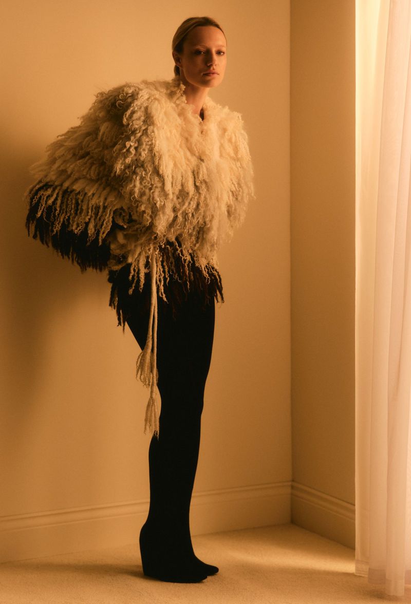 An adult woman poses in a textured fringe jacket, black leggings, and wedge heels indoors by a window with filtered light.