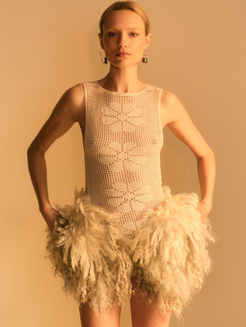 Woman in crochet bodysuit and fur shorts poses in front of a neutral background in a fashion-style studio shot with golden lighting.