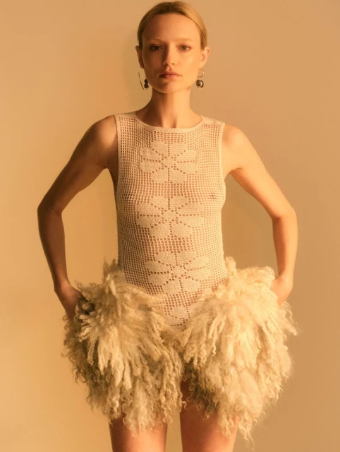 Woman in crochet bodysuit and fur shorts poses in front of a neutral background in a fashion-style studio shot with golden lighting.