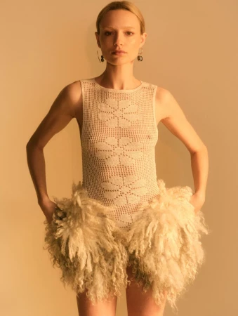 Woman in crochet bodysuit and fur shorts poses in front of a neutral background in a fashion-style studio shot with golden lighting.