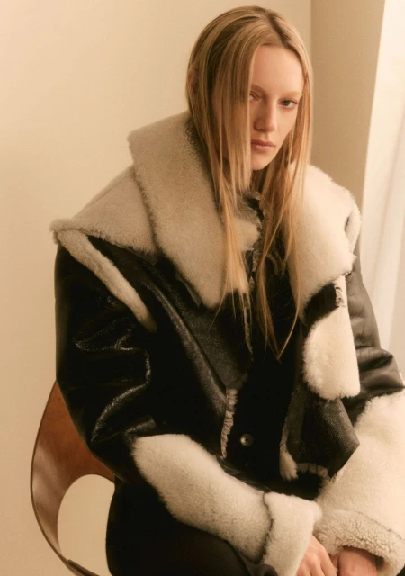 Blonde woman poses indoors wearing a black and white faux fur coat, seated on a brown chair in a fashion studio. Neutral background.