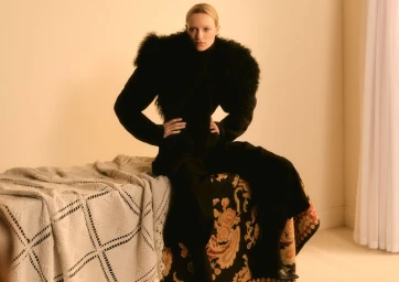 Model Maya Stepper wearing a black fur collared coat seated on a patterned quilt in a warmly lit room.