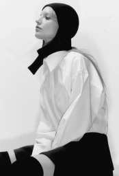 Model Maya Stepper wearing a black balaclava and an oversized white button-down shirt against a white studio background.