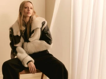 Model Maya Stepper wearing a black and white shearling jacket seated indoors near draped white curtains