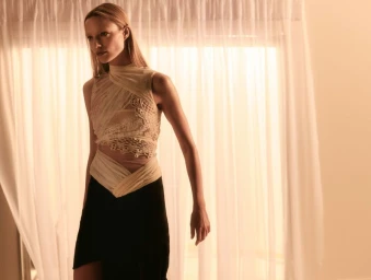 Model wearing a cream textured asymmetric sleeveless top and black skirt standing before sheer white curtains.