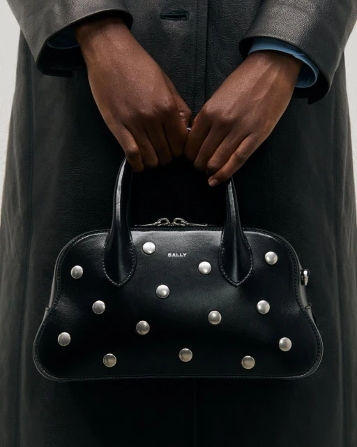 Woman in black coat holds a studded Bally handbag with a top handle against a neutral background, close up on the purse and hands.