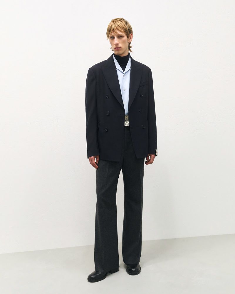 Man in a black double-breasted blazer and grey slacks stands in a white studio setting, creating a classic yet modern fashion statement.