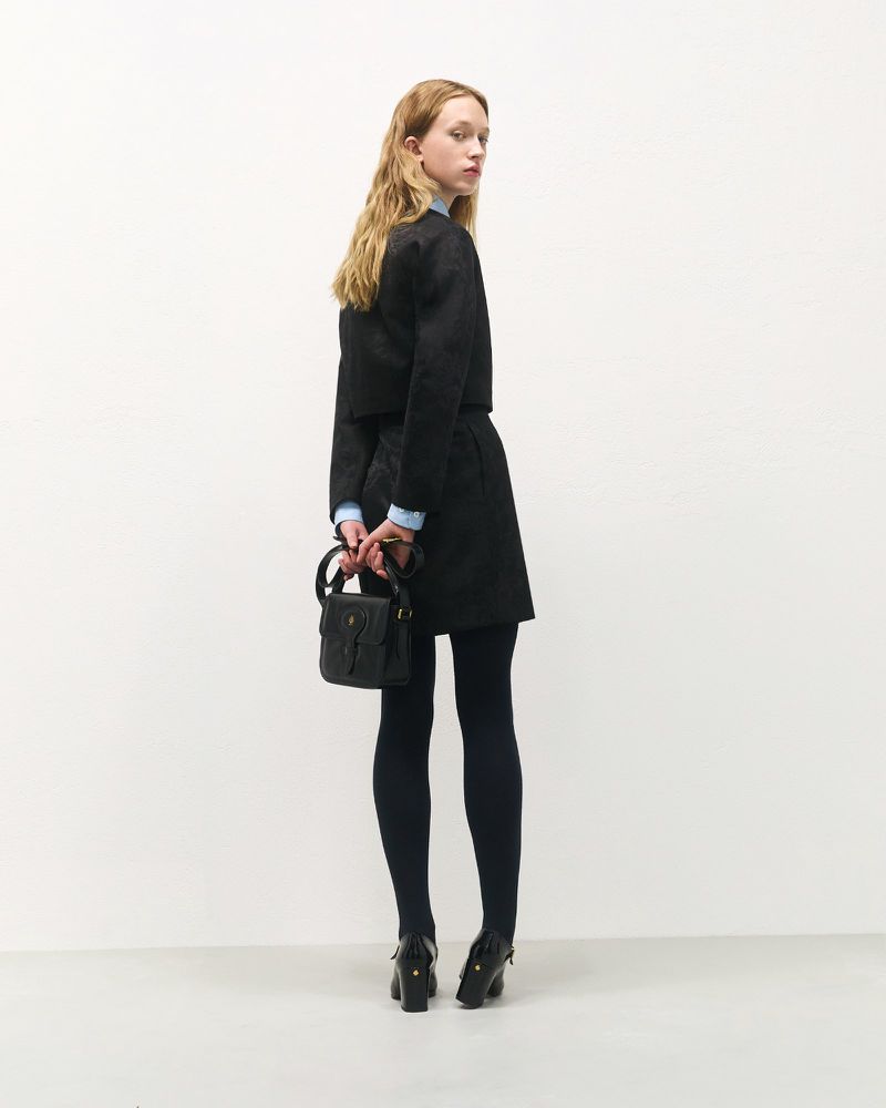 A woman stands with her back partially turned, wearing a dark skirt and jacket, complemented by navy tights and a black handbag.