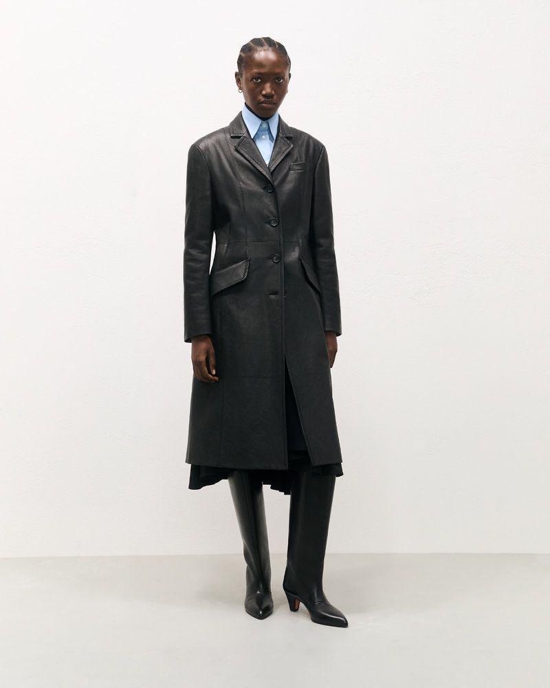 Elegant African woman poses in a black leather coat with a light blue shirt and black leather boots against a neutral backdrop.