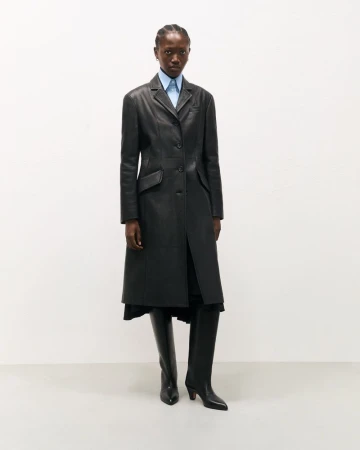 Elegant African woman poses in a black leather coat with a light blue shirt and black leather boots against a neutral backdrop.