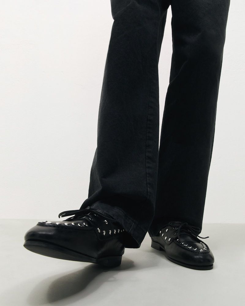 Black leather dress shoes with silver metal eyelets. The shoes are worn with dark trousers. The image is shot in a photo studio.