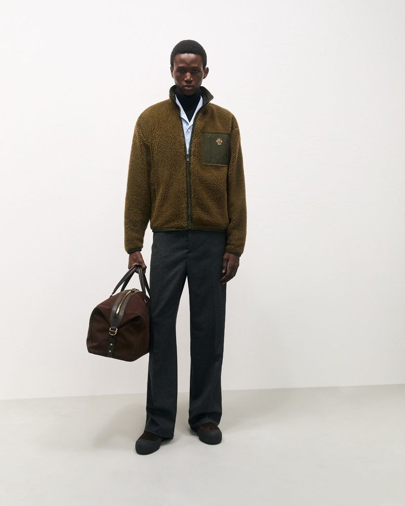 An adult male stands in a photo studio, wearing a sherpa jacket, dress pants, and carrying a duffel bag against a neutral background.
