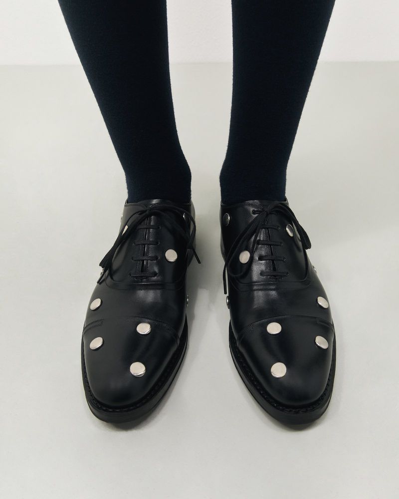 Elegant black leather Oxford shoes with silver button embellishments are paired with black tights in a minimalist setting. 
