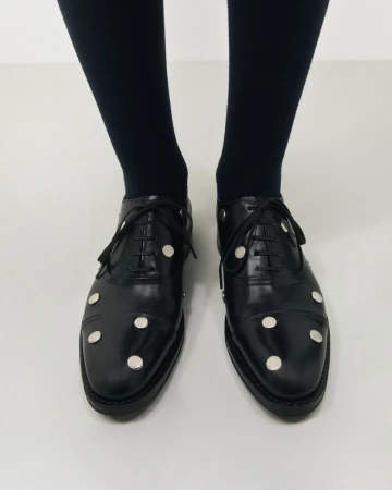 Elegant black leather Oxford shoes with silver button embellishments are paired with black tights in a minimalist setting. 