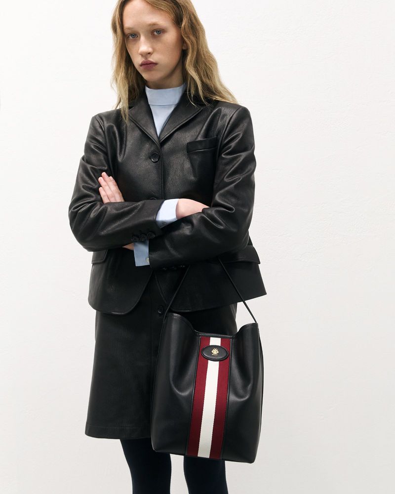 Woman in black leather suit with a black leather bucket bag featuring red and white stripes, standing against a neutral backdrop.