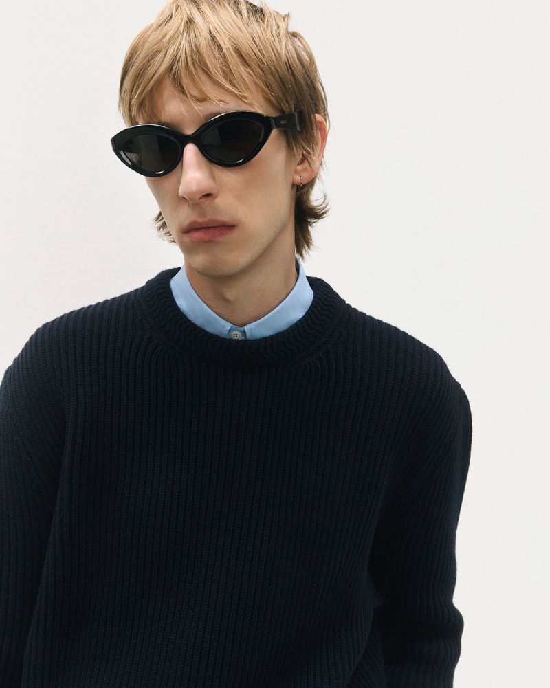 Close-up studio shot of a blonde adult man wearing sunglasses and a navy blue crewneck sweater against a white backdrop.