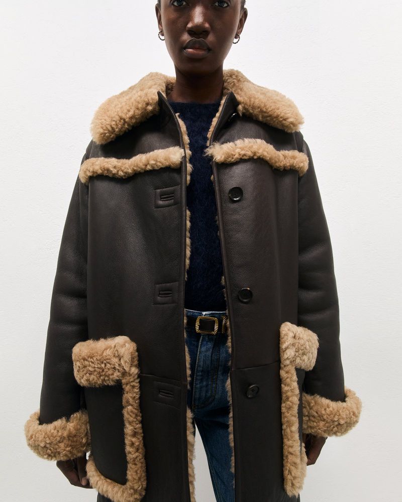 Dark-skinned woman wearing a brown shearling coat, posing in front of a plain white background in a fashion shot. Jeans and a sweater visible.