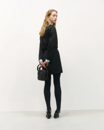 Model wearing dark jacket, matching skirt, and tights holding a black handbag against a plain white studio backdrop.