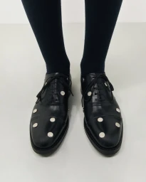 Close-up of black leather lace-up dress shoes embellished with round silver studs, worn with dark socks on a pale neutral surface.