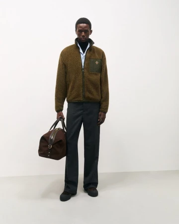 Model Gaye Serigne in an olive fleece zip jacket and dark trousers holding a brown duffle bag against a white studio background.