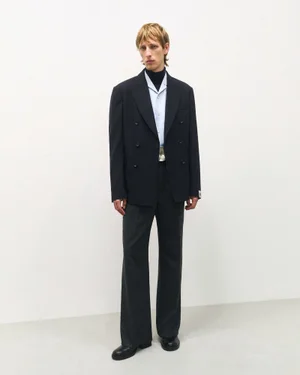Model wearing a dark double-breasted suit jacket over a light blue shirt and dark trousers against a white background