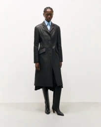Model wearing a tailored black leather coat, collared light blue shirt, and knee-high black boots against an off-white background.