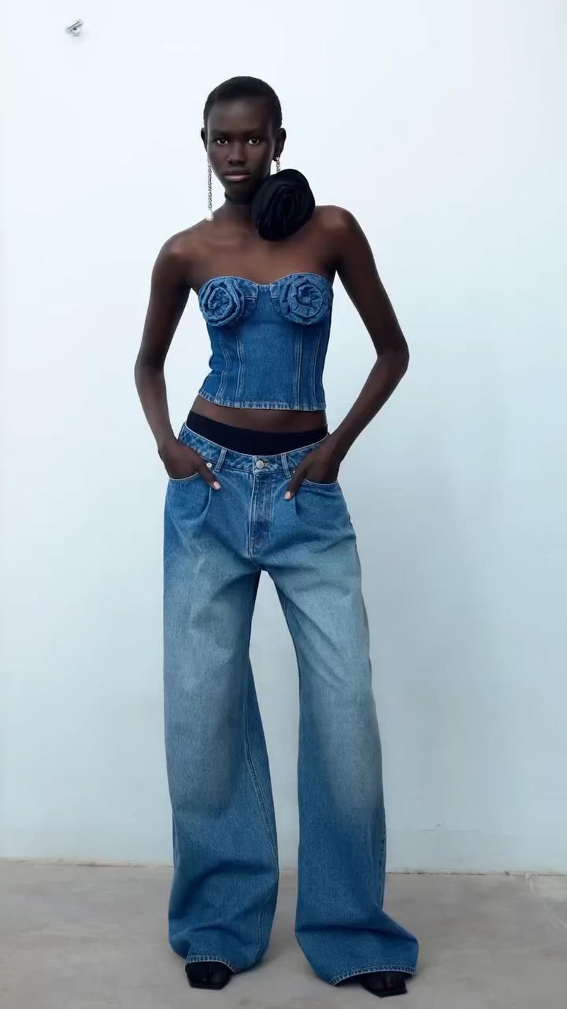 A slender woman with dark skin poses in a denim floral top and wide-leg jeans against a pale background in a fashion shot.