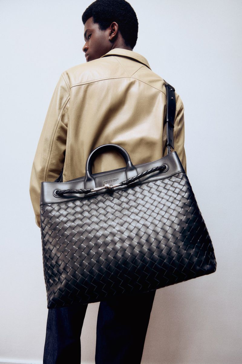 Model Willie Fidelis carrying a large black woven leather tote bag over his shoulder wearing a tan leather jacket against a white backdrop.