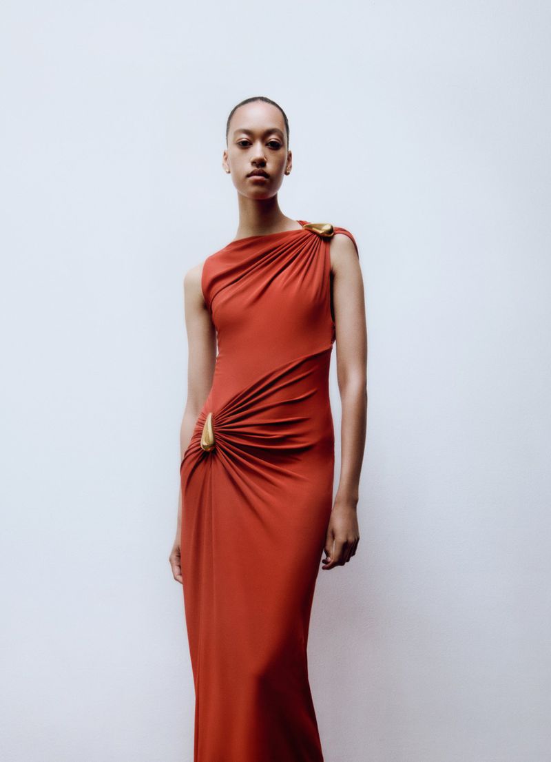 Elegant adult woman in a floor-length, rust-colored gown with draped fabric and gold clasps posing against a white background.