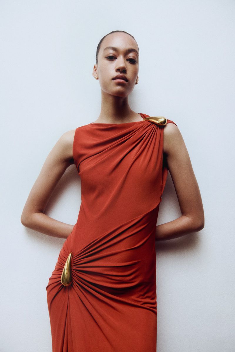 Elegant portrait of a woman in an orange sleeveless gown with draped fabric and gold accents posing against a stark white backdrop.