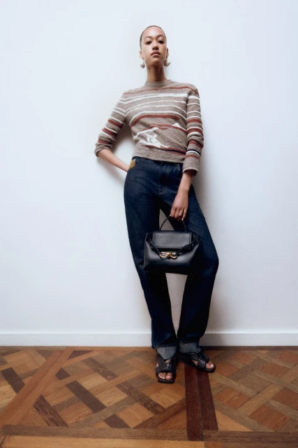 Full-shot fashion portrait of a woman posing in jeans, a sweater, and sandals, holding a black handbag in an indoor environment.