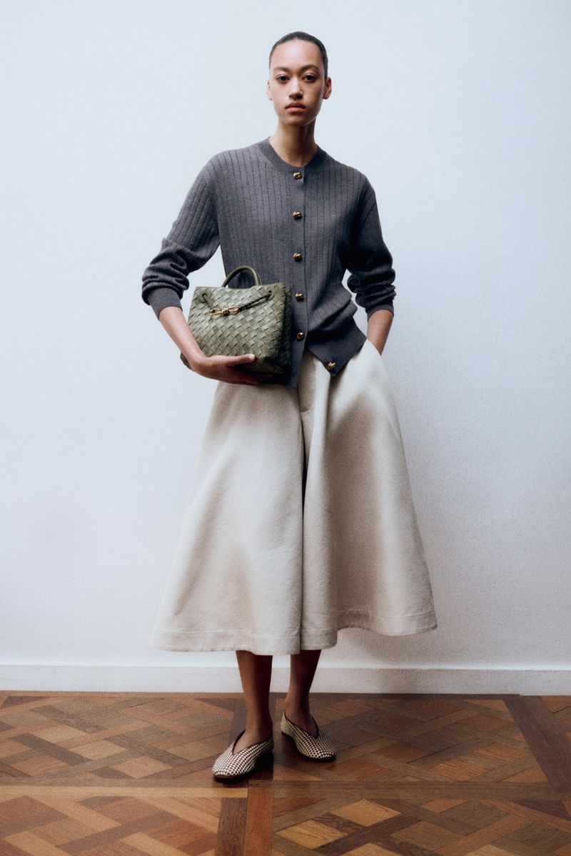 Elegant African American woman in grey cardigan, ecru culottes and woven flats, holding a woven handbag against a white wall.
