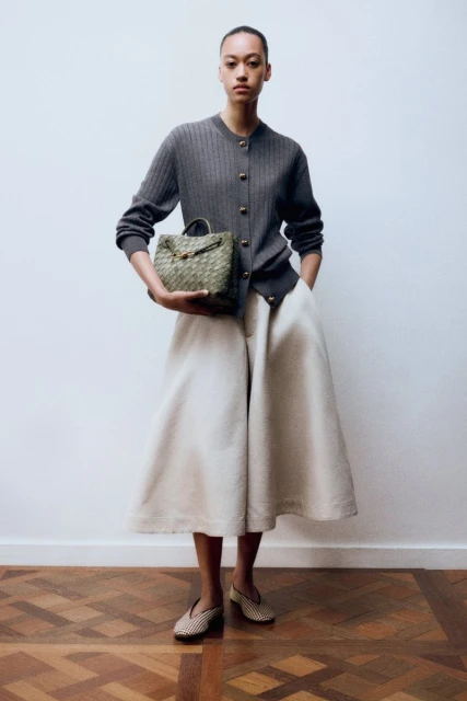 Elegant African American woman in grey cardigan, ecru culottes and woven flats, holding a woven handbag against a white wall.